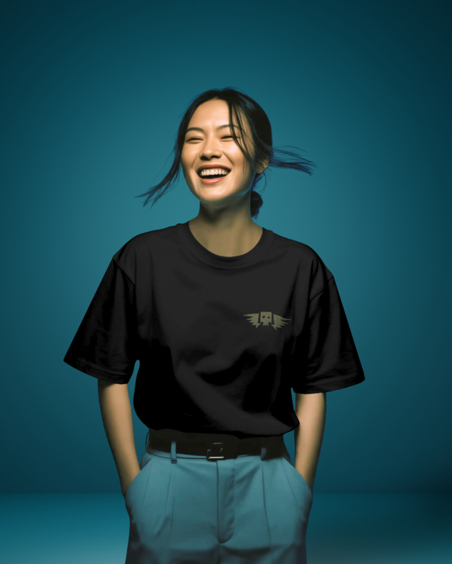Woman wearing Qurrito’s Black Winged Skull Oversized Streetwear Tee tucked into blue high-waist pants. The subtle embroidery and dark tone project confidence and mystery mood, creating a bold yet minimalist streetwear outfit.