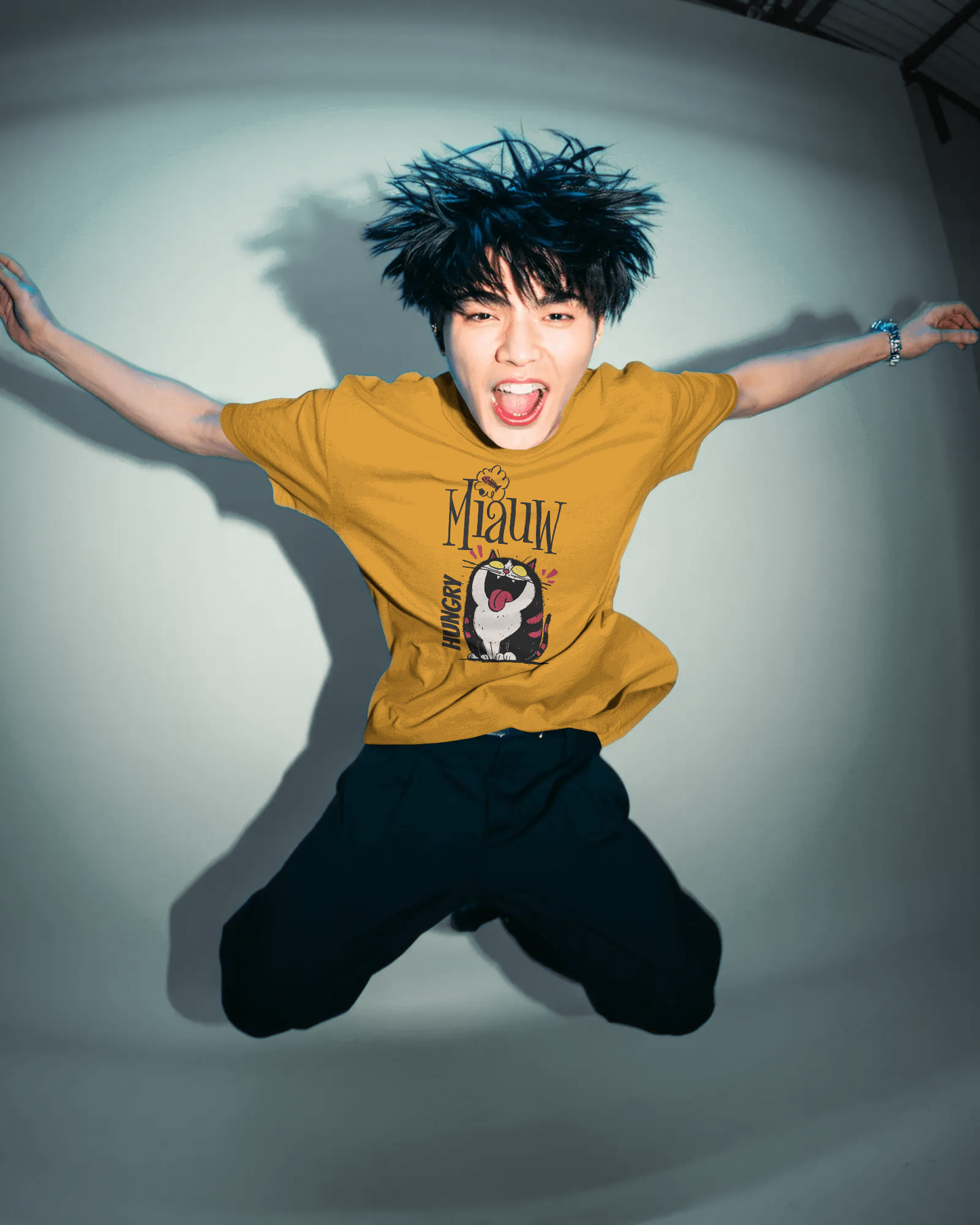 Qurrito Level 100 Hangry graphic t-shirt in Regular Fit worn by a man jumping in an energetic pose. The mustard yellow tee features the Energetic Funny Graphic of a black and white cat yelling "Miauw" and "HUNGRY." Energetic mood.