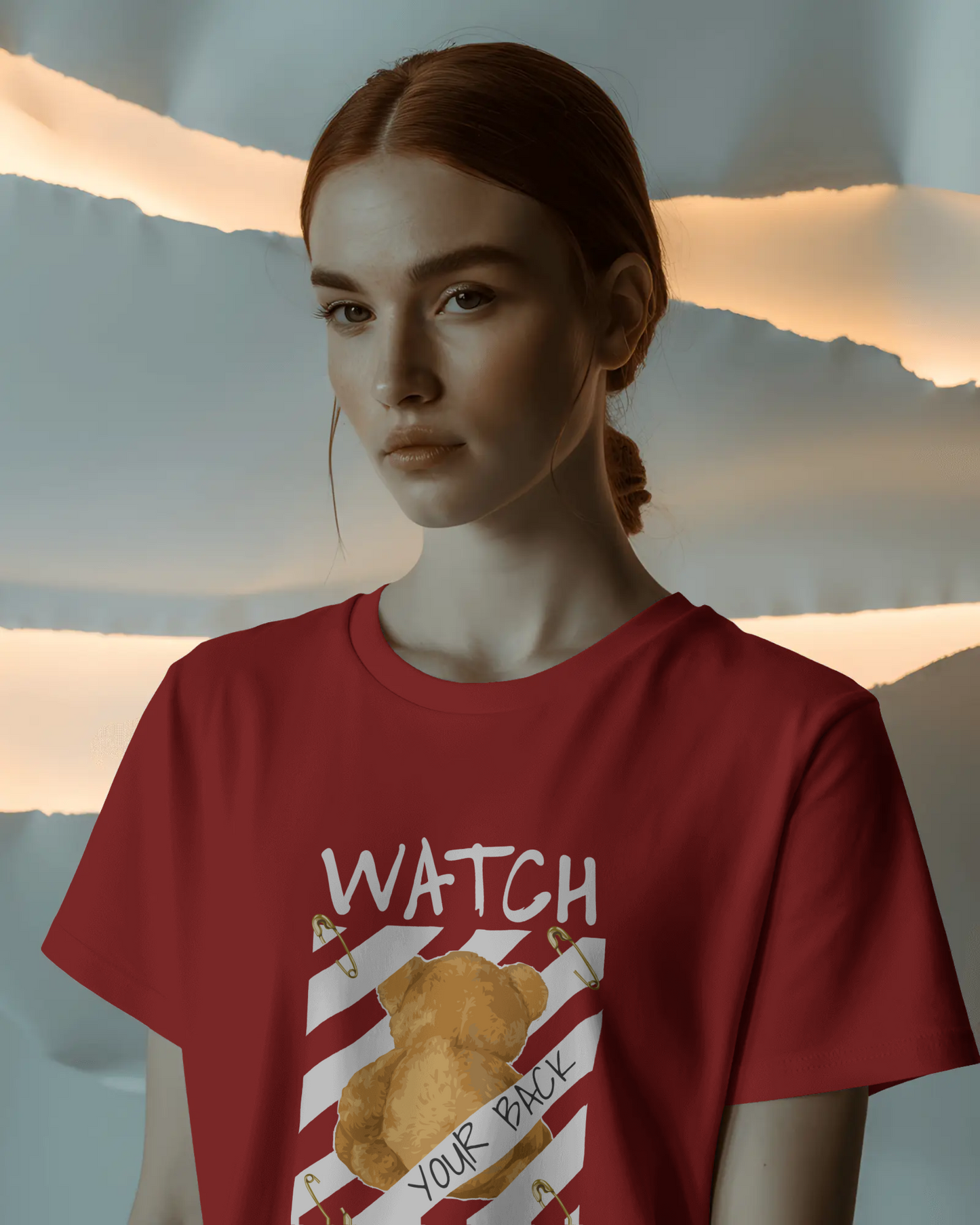 Red oversized t-shirt featuring a teddy bear graphic facing away with the bold text 'Watch Your Back,' safety pin details, and diagonal caution stripes. Includes the phrase 'Real world is behind your back' at the bottom. Urban graphic wear style by Qurrito.