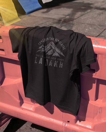 Qurrito’s Mountain Graphic Ladakh T-Shirt draped over a barrier under sunlight, showcasing “Conquer the Himalayas – The Land of High Passes” print in coffee brown. This travel-themed regular fit tee expresses serious, focused, chill, relaxed, happy, and cheerful moods — perfect for adventure and mountain lovers.