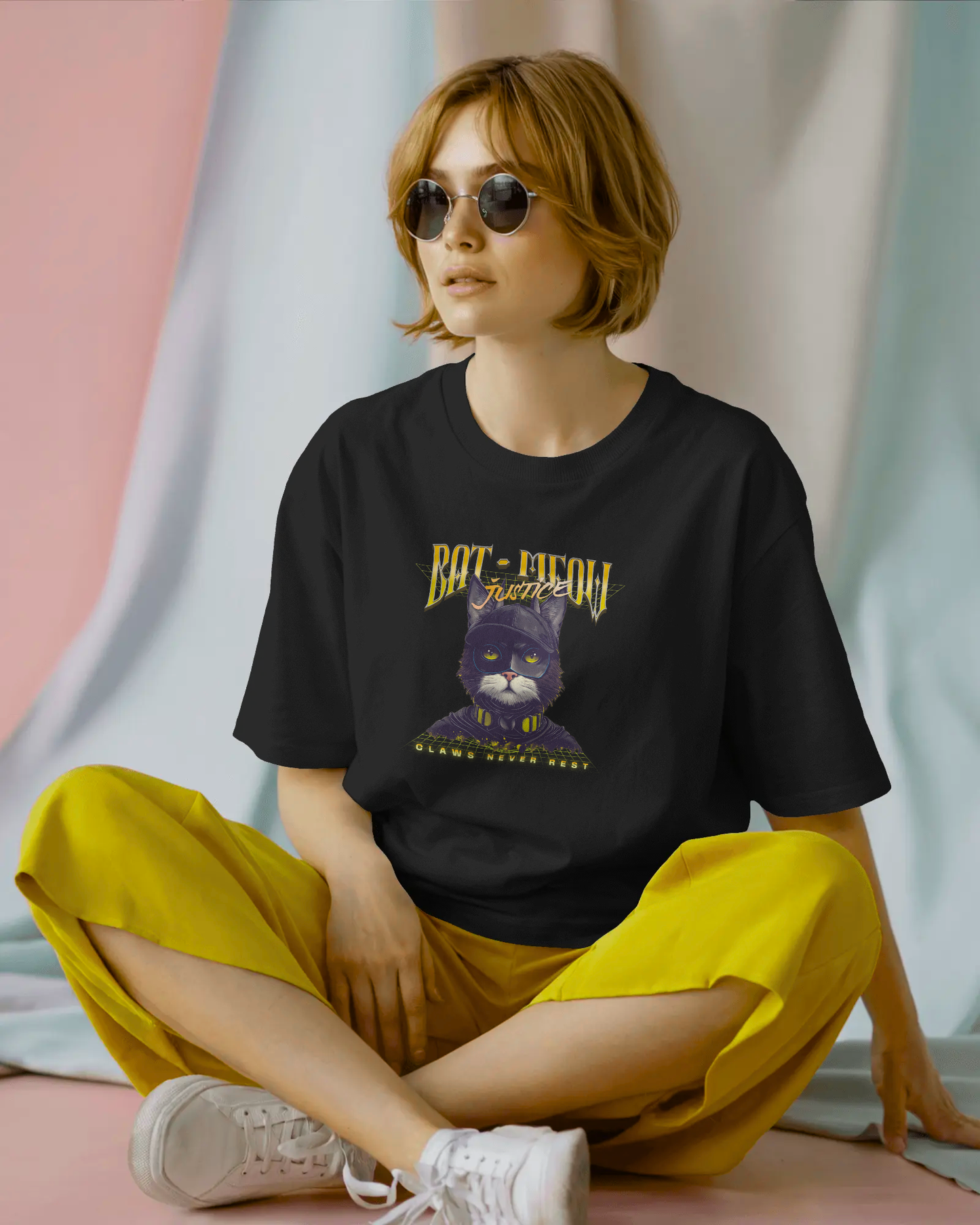 Playful and quirky female model wearing a black oversized unisex T-shirt featuring the "Bat-Meow Justice" superhero cat graphic. The tee has a drop-shoulder fit and is styled with bright yellow pants and sunglasses for a fun look.