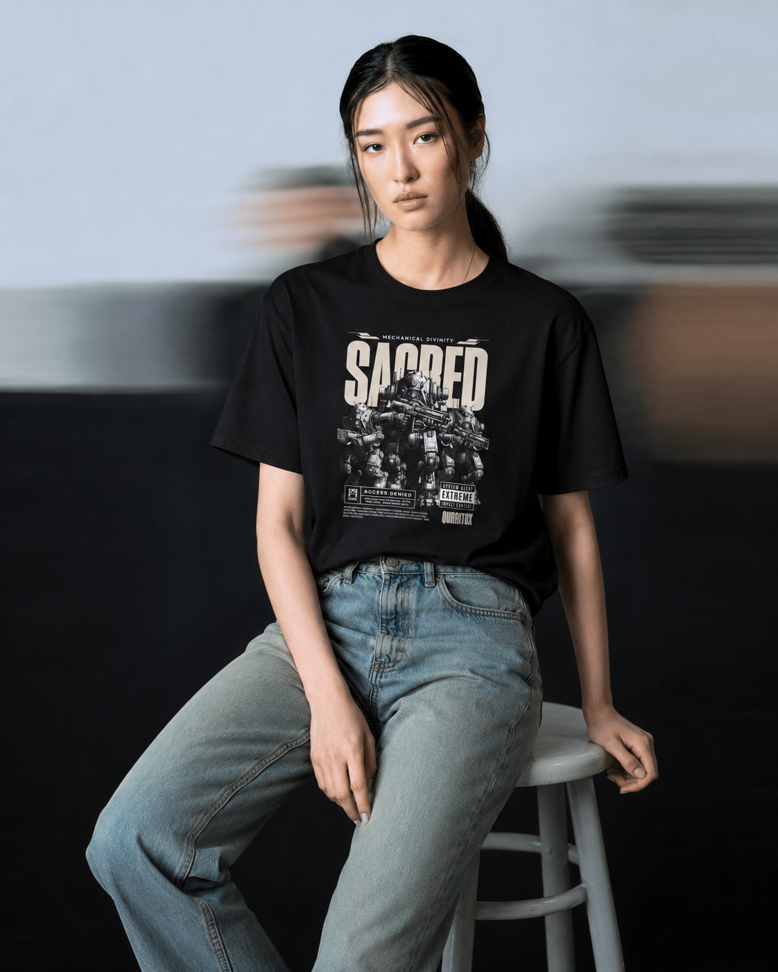 Woman sitting on stool wearing QURRiTO Sacred Mecha Divinity black cyberpunk t-shirt one size up for an oversized vibe, paired with light wash jeans in a dark studio setting.
